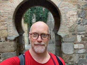 La imagen muestra a un hombre con camisa roja frente a un muro de ladrillo arqueado en el que crece vegetación, posiblemente tomándose un selfie.