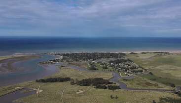 This image shows a coastal town bordered by the sea and surrounded by green fields, winding roads, a river, and patches of vegetation under a partly cloudy sky.