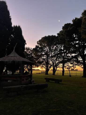 A peaceful rural scene at sunset, with silhouettes of trees, wooden benches, a thatched hut, a crescent moon, and fields stretching into the horizon.