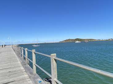 Un muelle de madera se adentra en las aguas azules, con barandillas y farolas. Varias embarcaciones flotan cerca. Al fondo se ve una pequeña colina y un cielo despejado.