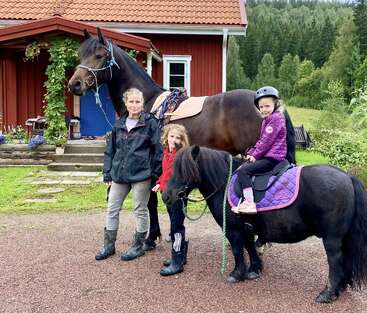 Une femme et deux enfants posent à l'extérieur, près d'une maison. L'un des enfants est assis sur un petit poney, tandis que la femme tient un cheval par la bride. Ils sont entourés de verdure.