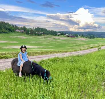 Une fillette souriante, vêtue d'une robe bleue et coiffée d'un casque, est assise sur un petit poney noir qui broute dans une prairie verdoyante, avec en arrière-plan des terres agricoles pittoresques et des nuages spectaculaires.