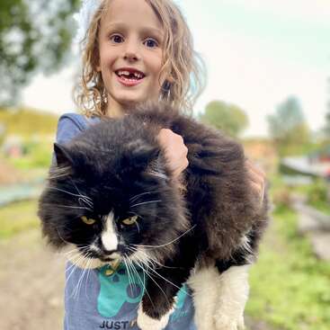 Un enfant joyeux, à qui il manque une dent, tient dans ses bras, à l'extérieur, un gros chat noir et blanc au pelage soyeux, et sourit de toutes ses dents. Le chat a l'air détendu et à l'aise dans ses bras.