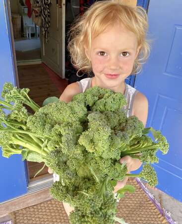 Un jeune enfant aux cheveux blonds, tout sourire, se tient dans l'embrasure d'une porte, tenant à deux mains un gros bouquet de brocolis fraîchement cueillis, qu'il présente fièrement.