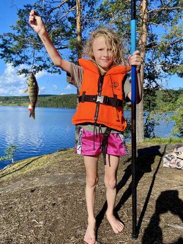 Au bord d'un lac pittoresque, entouré d'arbres et baigné de soleil, un jeune enfant vêtu d'un gilet de sauvetage orange tient fièrement une canne à pêche et un poisson fraîchement pêché.