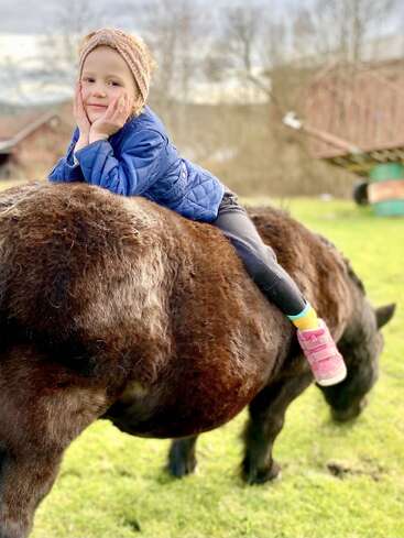Un jeune enfant, vêtu d'une veste bleue et de chaussures roses, est allongé, l'air heureux, sur un poney brun dans un décor extérieur verdoyant ; il sourit, les mains posées sur les joues.
