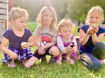 Quatre jeunes enfants sont assis dans l'herbe, baignés par la lumière du soleil, chacun tenant un petit poussin dans ses bras. Ils sourient et bercent délicatement les poussins, profitant d'un moment paisible et joyeux en plein air.