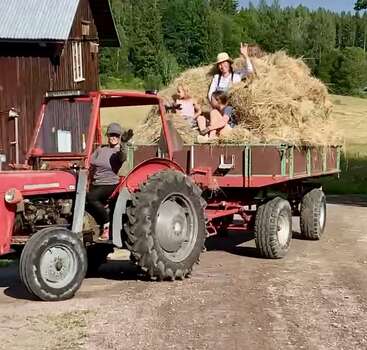 Un tracteur rouge tire une remorque remplie de foin, tandis que des gens souriants saluent de la main. Ils se trouvent en plein air, près d'une grange rustique, avec des arbres verdoyants en arrière-plan.