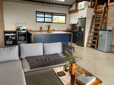 Ein moderner, gemütlicher, offener Küchen- und Wohnbereich mit einem grauen Sofa, einem Couchtisch aus Holz, einer Loftleiter, Edelstahlgeräten und natürlichem Licht durch große Fenster.