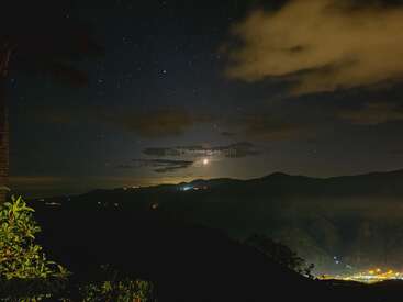Ein atemberaubender Nachthimmel, gefüllt mit unzähligen Sternen, erhebt sich über dunklen Bergen. Die Wolken schweben sanft, die Lichter der Stadt leuchten darunter und das Laub rahmt die ruhige, mondbeschienene Landschaft ein.