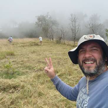 Un homme souriant portant un chapeau prend un selfie, affichant un signe de paix dans un champ brumeux et herbeux, tandis que deux personnes marchent devant lui parmi des arbres clairsemés.