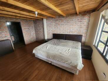 Esta habitación cuenta con techo de madera con vigas vistas, paredes de ladrillo, una cama grande, dos mesitas de noche, suelo de madera, una ventana y una puerta abierta al cuarto de baño.