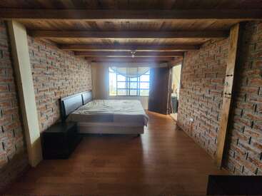 Esta habitación rústica cuenta con paredes de ladrillo visto, vigas de madera en el techo y suelo de madera noble. La cama y la mesita de noche están situadas frente a una ventana luminosa con luz natural.