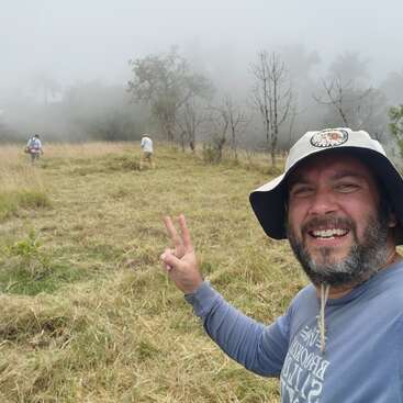 A smiling man wearing a hat takes a selfie, flashing a peace sign in a misty, grassy field while two people walk ahead among sparse trees.
