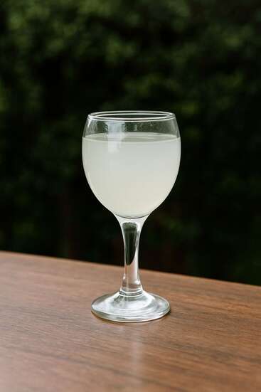 Un verre à vin transparent est posé sur une table en bois, rempli d'un liquide pâle et trouble. L'arrière-plan est constitué d'un feuillage vert flou, créant un environnement paisible en plein air.