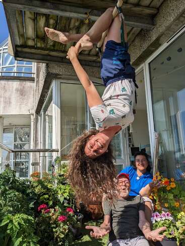 A woman hangs upside down, smiling joyfully, while two people laugh below her among vibrant flowers on a sunny balcony, radiating fun, happiness, and togetherness.