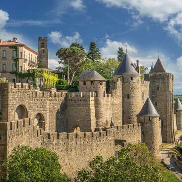 This image shows an ancient stone castle with tall towers and crenellated walls, surrounded by lush greenery, under a bright blue sky with clouds.