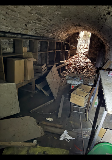 Dieses Bild zeigt einen vollgestopften, staubigen Steinkeller mit Kisten, alten Holzmöbeln, Regalen und Gerümpel, beleuchtet von schwachem Tageslicht, das durch einen Tunneleingang hereinfällt.
