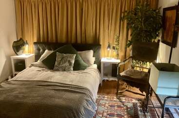 Ein gemütliches Schlafzimmer mit einem großen Bett, gemusterten Kissen, Nachttischen mit Lampen, einem Holzstuhl, einer großen Pflanze, goldenen Vorhängen und einem dekorativen Teppich.