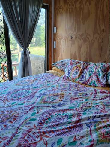A cozy bedroom with colorful, patterned bedding, yellow pillows, wooden walls, and a large window with sheer gray curtains overlooking a green, sunny outdoor area.
