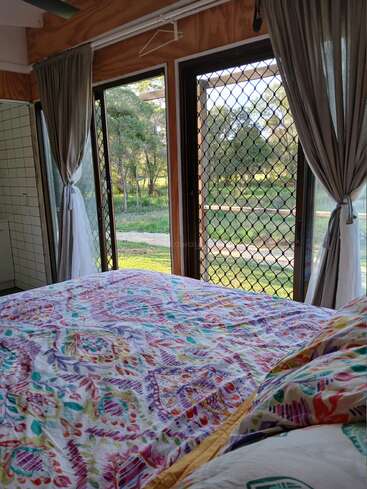 A cozy bedroom with colorful, patterned bedding faces large windows and a screen door, offering a scenic view of green trees, sunlight, and peaceful nature outside.