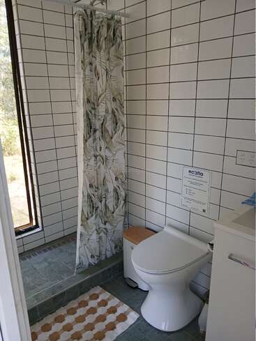 This bathroom features white tiled walls, a green tiled floor, a shower with a leafy curtain, a modern toilet, vanity, window, and a dotted mat.
