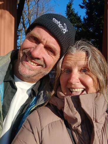 Un homme souriant portant un bonnet noir et une femme joyeuse portant une veste marron posent pour un selfie en plein air par une journée ensoleillée et claire.