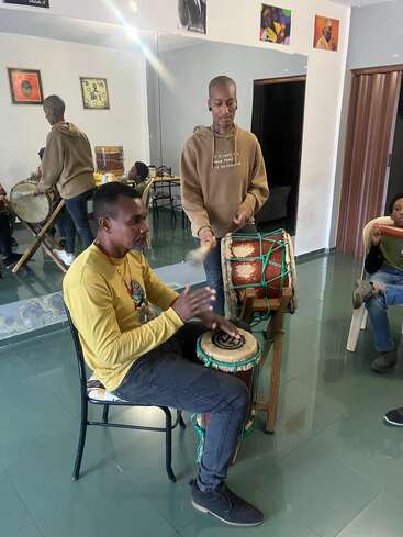 Two people play traditional African drums in a bright, art-filled room. Others observe, creating a lively, communal atmosphere focused on music and cultural connection.