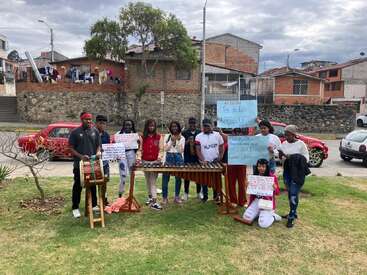 A group of young people stands outdoors with a marimba and drum, holding handwritten signs, protesting or celebrating Afro culture in a residential neighborhood.