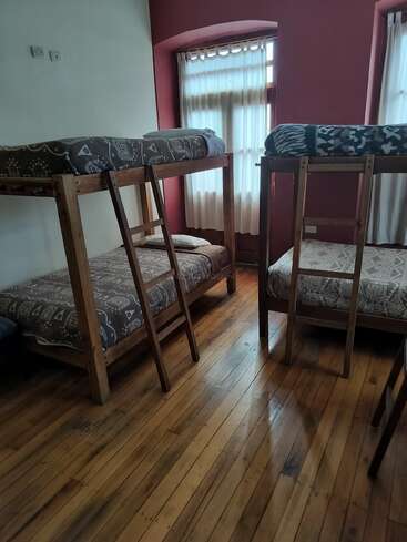 This image shows a cozy room with wooden floors, two sets of sturdy bunk beds, patterned bedding, soft lighting, and large windows with sheer curtains.