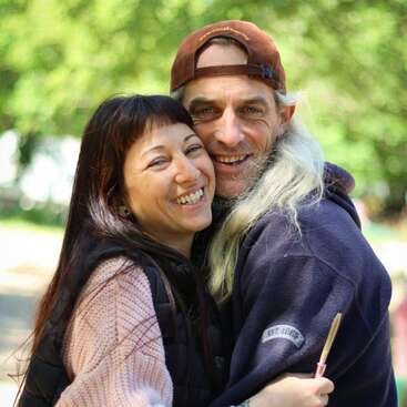 Um casal feliz se abraça ao ar livre, sorrindo calorosamente para a câmera. O fundo é verde e exuberante. Eles usam jaquetas aconchegantes, irradiando afeto e alegria juntos.