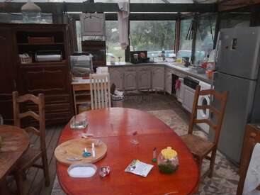 La imagen muestra una cocina rústica con una mesa ovalada roja, sillas de madera, un frigorífico grande, armarios y electrodomésticos. La luz natural entra por grandes ventanales, mostrando verdor en el exterior.