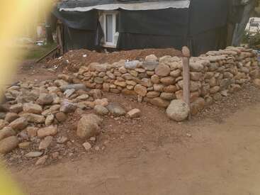 Un mur de pierre rustique, partiellement construit, entoure une zone en terre battue à côté d'une structure recouverte d'un tissu noir. Des pierres éparpillées jonchent le sol à proximité, et un poteau en bois est planté verticalement.