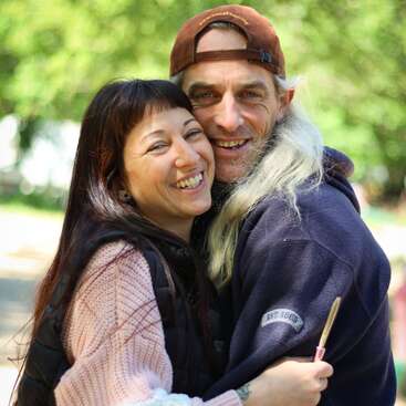 Una pareja feliz se abraza al aire libre, sonriendo cariñosamente a la cámara. El fondo es exuberante y verde. Llevan chaquetas acogedoras, irradiando afecto y alegría juntos.