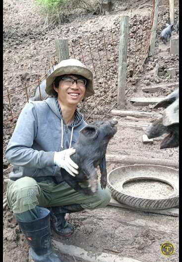 A smiling person wearing glasses, a hat, gloves, and boots is holding a small black piglet in a muddy outdoor farm area near a feeding trough.
