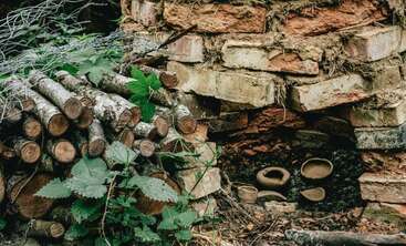 A rustic outdoor brick kiln with pottery inside. Stacked firewood and green leafy plants are nearby, surrounded by wire, creating a natural, earthy, and primitive scene.