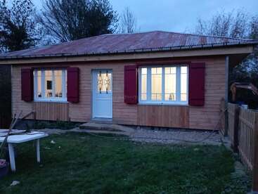 A small wooden house with a metal roof and red shutters, warmly lit from inside. It is surrounded by a grassy yard, trees, and a wooden fence.