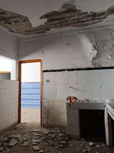 This image shows a dilapidated room with collapsed ceiling plaster, broken tiles, debris scattered on the floor, and a partially visible adjacent room with blue patterned tiles.
