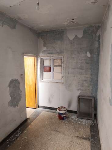 Essa foto mostra um cômodo pequeno e abandonado, com a pintura descascando, uma janela velha fechada, um balde de tinta no chão e alguns detritos de obra espalhados pelo chão.