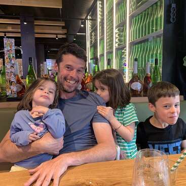 Das Bild zeigt einen Mann, der mit drei Kindern an einem Tisch sitzt, vor einer Bar mit Alkoholflaschen und einer grünen Wand mit Lichtern hinter ihnen.