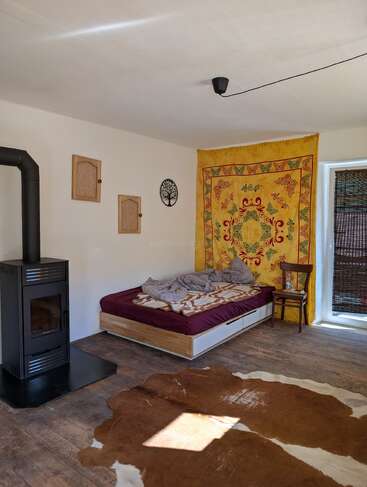 A cozy room with a wood stove, bed, and colorful tapestry. Wooden floor, animal hide rug, simple wall art, chair, and natural light create a warm atmosphere.