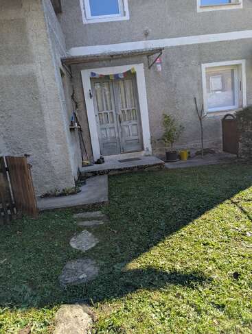 An old rustic house entrance with double doors, colorful prayer flags, concrete path, green grass, potted plants, and sunlight casting shadows across the yard.