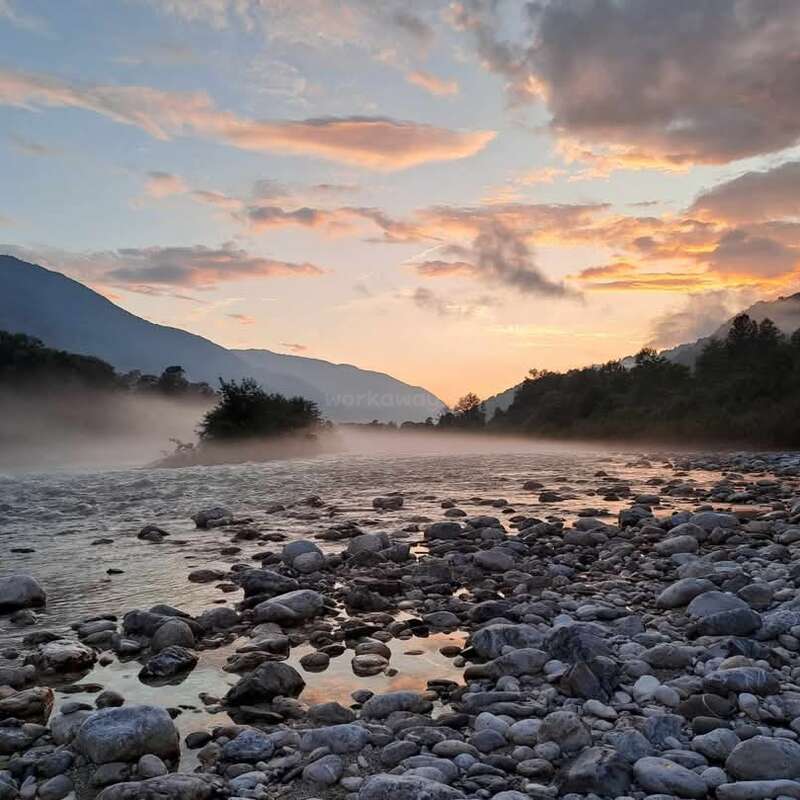 The image depicts a serene river scene at sunset, featuring a rocky shoreline, misty water, and distant mountains, set against a vibrant sky with pink and orange hues.