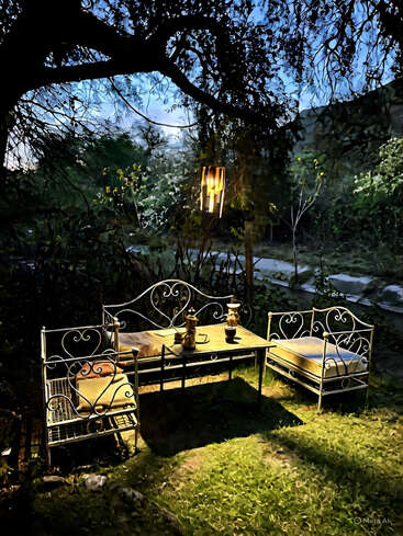 An inviting outdoor seating area with ornate wrought iron benches and table is softly lit under a tree. Evening ambiance is enhanced by warm lantern light.