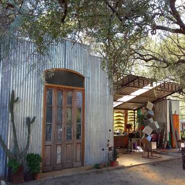 A rustic metal building with wooden doors stands beneath tree branches. An art studio or workshop, visible inside, displays creative sculptures, tools, and colorful materials.