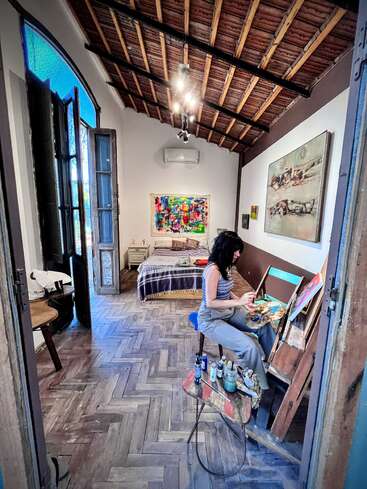 A woman paints at an easel in a cozy, artistic bedroom with high wooden ceiling, expressive artwork, wooden floors, and natural light streaming through open windows.