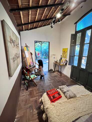 A cozy art studio bedroom with a wooden ceiling, bed, and blankets. A person paints at an easel. Natural light enters from open doors.