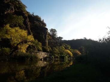 A serene river reflects lush green trees and a rocky hillside, bathed in soft sunlight. A stone bridge crosses the water, blending into nature’s tranquility.