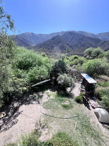 A sunlit mountainous landscape with dense green foliage, a dirt yard, scattered garden tools, and a shed. Towering mountains rise majestically in the background under clear skies.