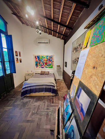 This cozy room features a single bed, wooden ceiling, blue window, various colorful artworks, an easel, and warm lighting, creating a creative and inviting atmosphere.
